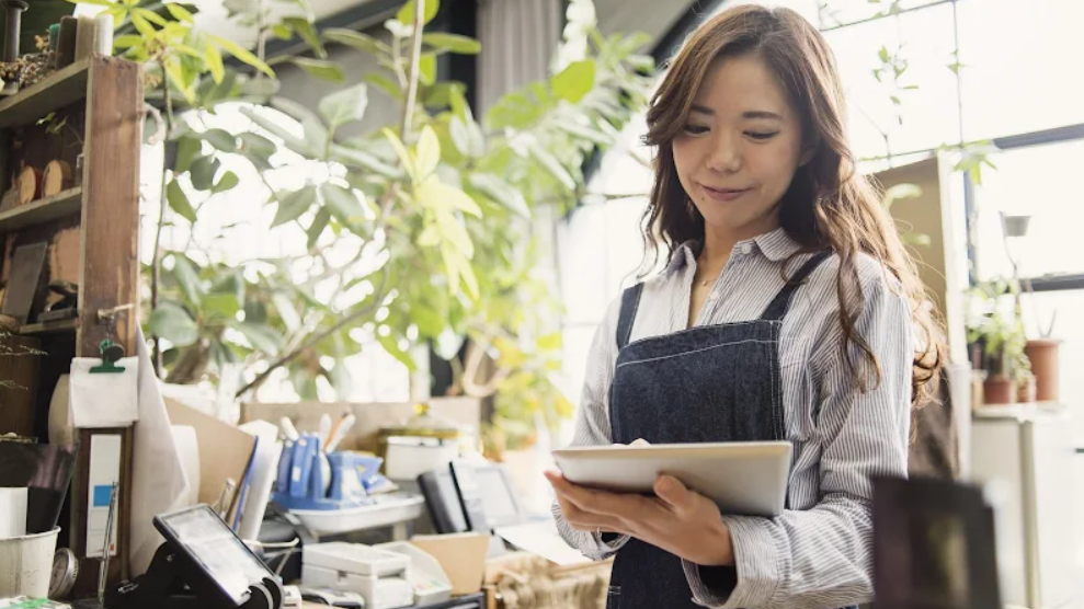 A woman examines a tablet while standing in a plant-filled office with a desk and office supplies.